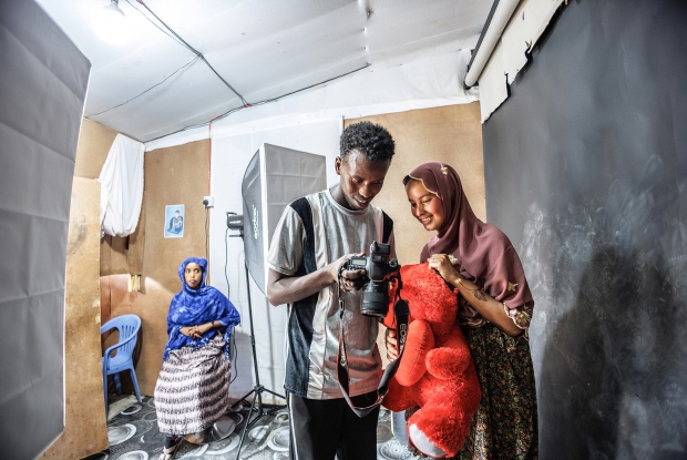 May 11th, 2024- Sheder Refugee Camp, Northeastern Fafan Zone in the Somali region of Ethiopia. Khadar interacts with a client during a photo session at his photo studio. &ldquo;My parents say that the major reason as to why they left Somalia was because of how compromised our safety had become. I have since been staying in this Refugee Camp with my family of 16. I started and finished my studies here. Right from primary to secondary school and finally, the university studies here. Now, I own this photo studio.  In this refugee camp setting, I feel like life keeps getting harder to maneuver financially, but I am glad that the idea of starting up this studio came to my mind. It has made it easier for me to contribute to my family's income. Before the solar power project came here, there was a big challenge in accessing electricity. We would pay for electricity per month but they would only switch it on for 3 hours a day. They would switch it on at 6:30 or 6:40 in the evening and then switch off at exactly 10. This is distasteful for any business that's dependent on electricity. People in this community had to go to neighboring towns where photo studios had access to electricity for services as basic as a passport photo. My business never made sense until when I knew about the plans to have us connected to a reliable source of power and since the connection, the problem has gotten behind me. People can now access my services all day and I am also making some good money. The money that I used to make in 4 days, I now make it in 1 day.