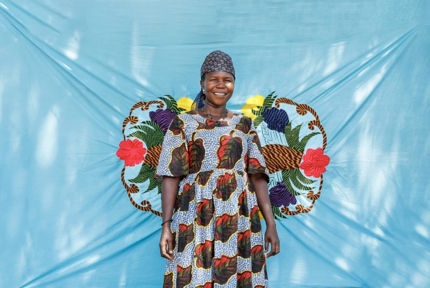 07/06/2024, Bidibidi Refugee settlement, Uganda. Rahima Yusuf standing in front of one of her custom-designed bedsheets. She buys the plain cotton material and knits designs into it, to make a more appealing bedsheet that she sells at a profit. Upon her arrival at the camp, Rahima, a Single mother of five children relied on humanitarian relief assistance provided by the World Food Program and UN Agencies in the camp and it was hard for her to provide all the family basics. The DREAMS program provided Rahima with a seed grant, enabling her to start a business retailing mixed goods. Whenever Rahima has a minute to spare, she makes/designs bedsheets for sale. Her current business value is worth about 1.6 million Uganda shillings. These enterprises enabled her to support her children by paying their school requirements, medical bills, clothing, and food.